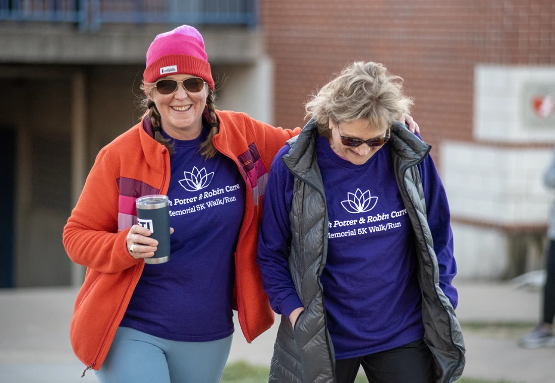 Two Knox alumni wearing Knox t-shirts laugh and walk, arm in arm, on campus.