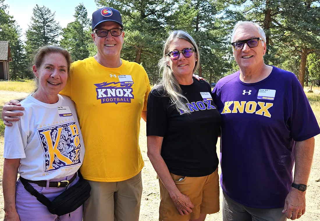 Four Knox alumni in Prairie Fire gear gather and smile at the camera.