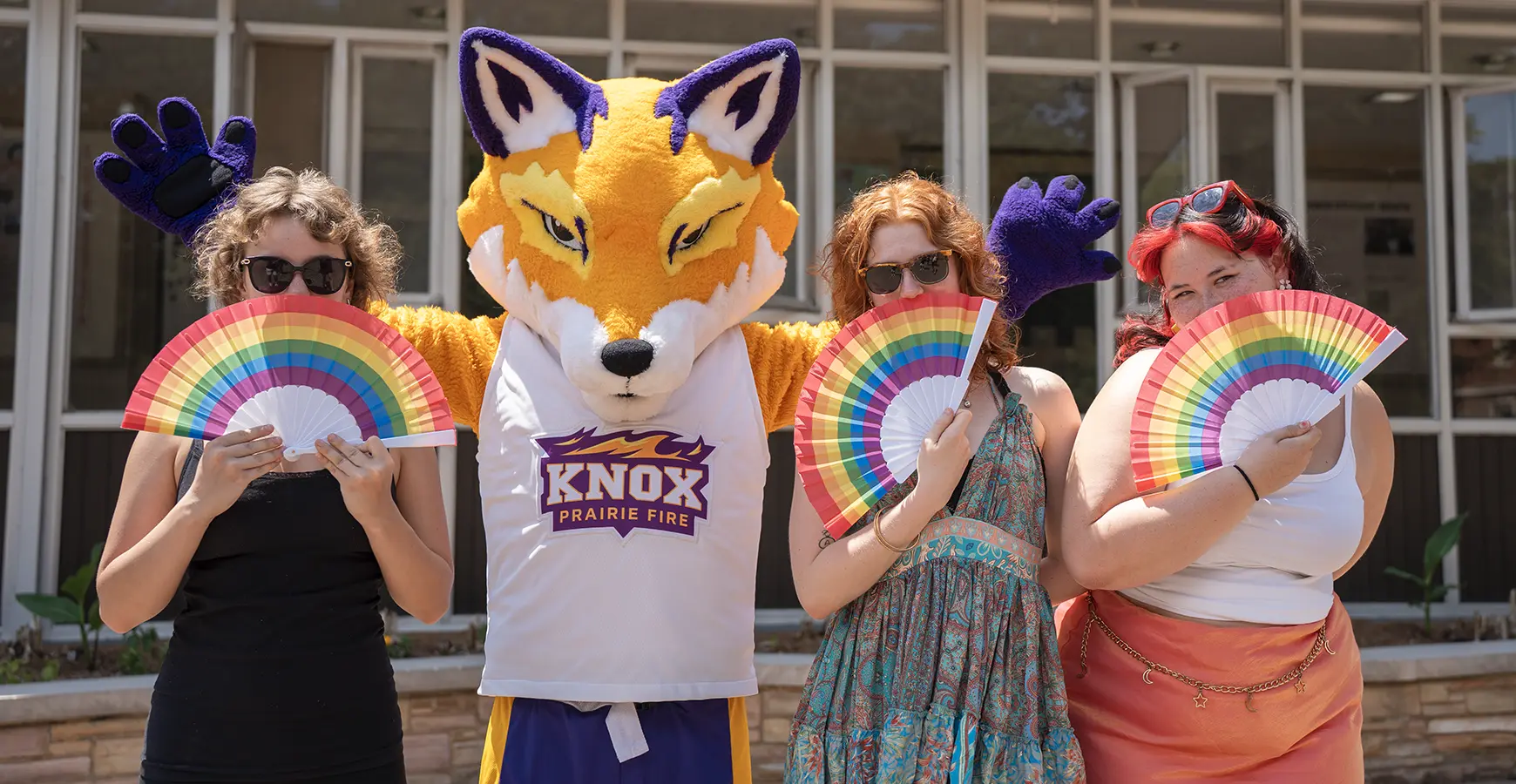 Three students stand with Blaze the mascot and hold up rainbow fans over their faces.