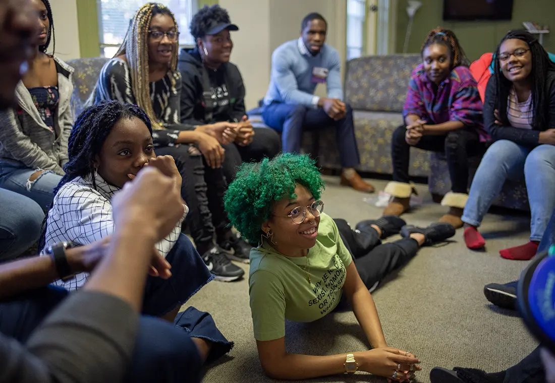 A group of students sits on the floor and furniture, smiling while listening to an alum speak.