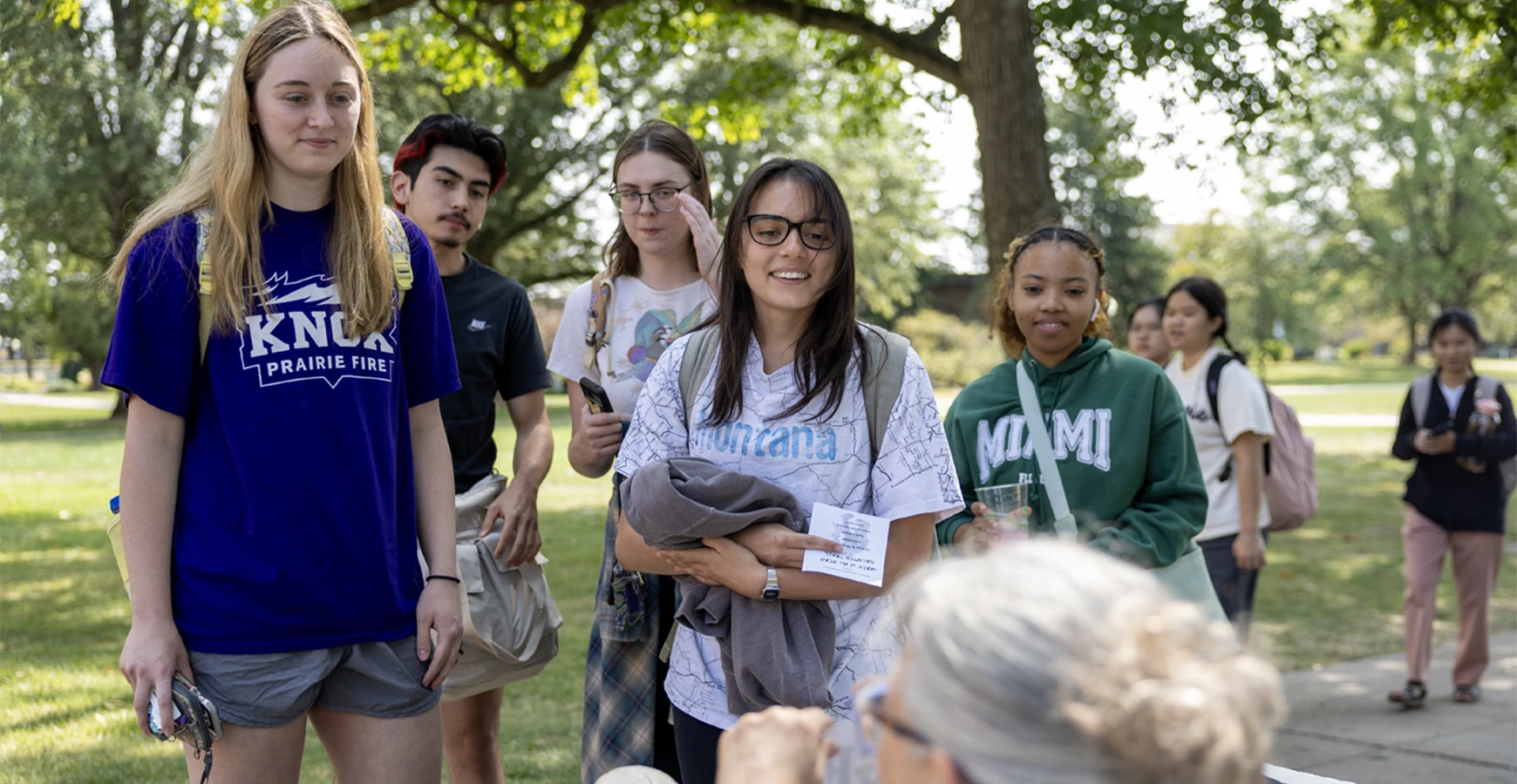 A group of students stand in front of a person sitting at a table, smiling at them.