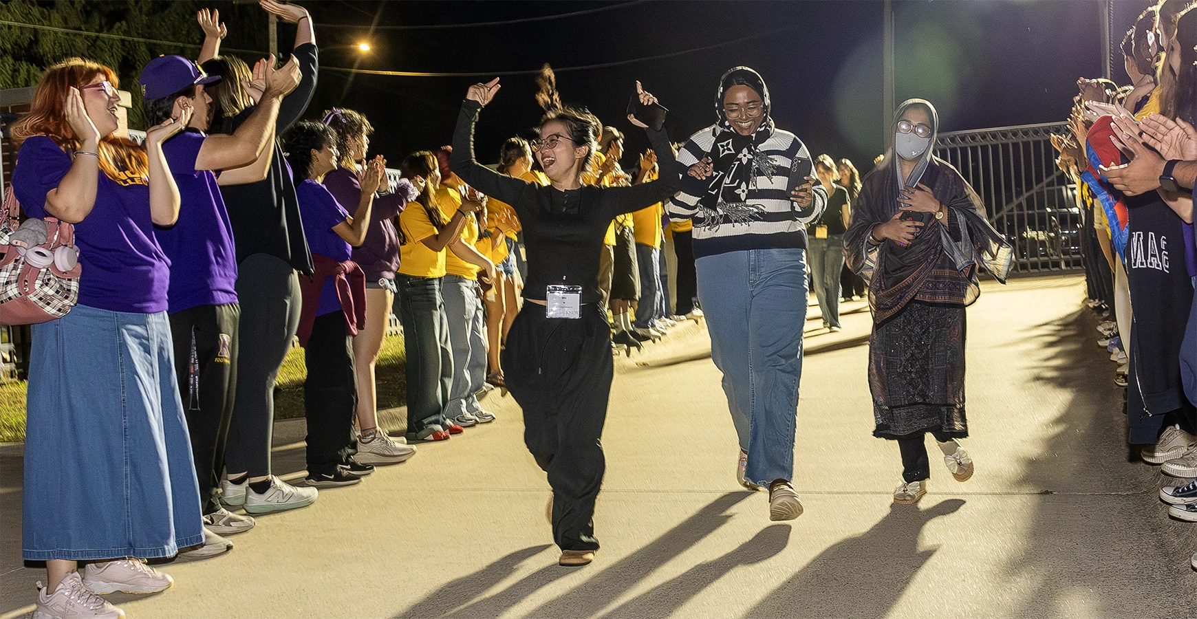 Three students walk down a sidewalk surrounded by other students cheering for them.