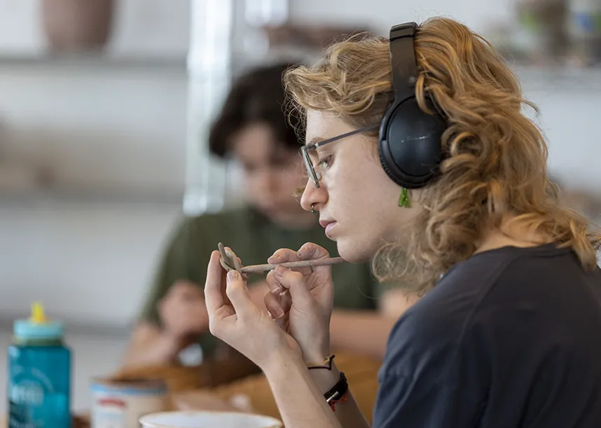 A student wearing headphones holds a small piece of clay in one hand and uses a scraping tool on the clay with their other hand.