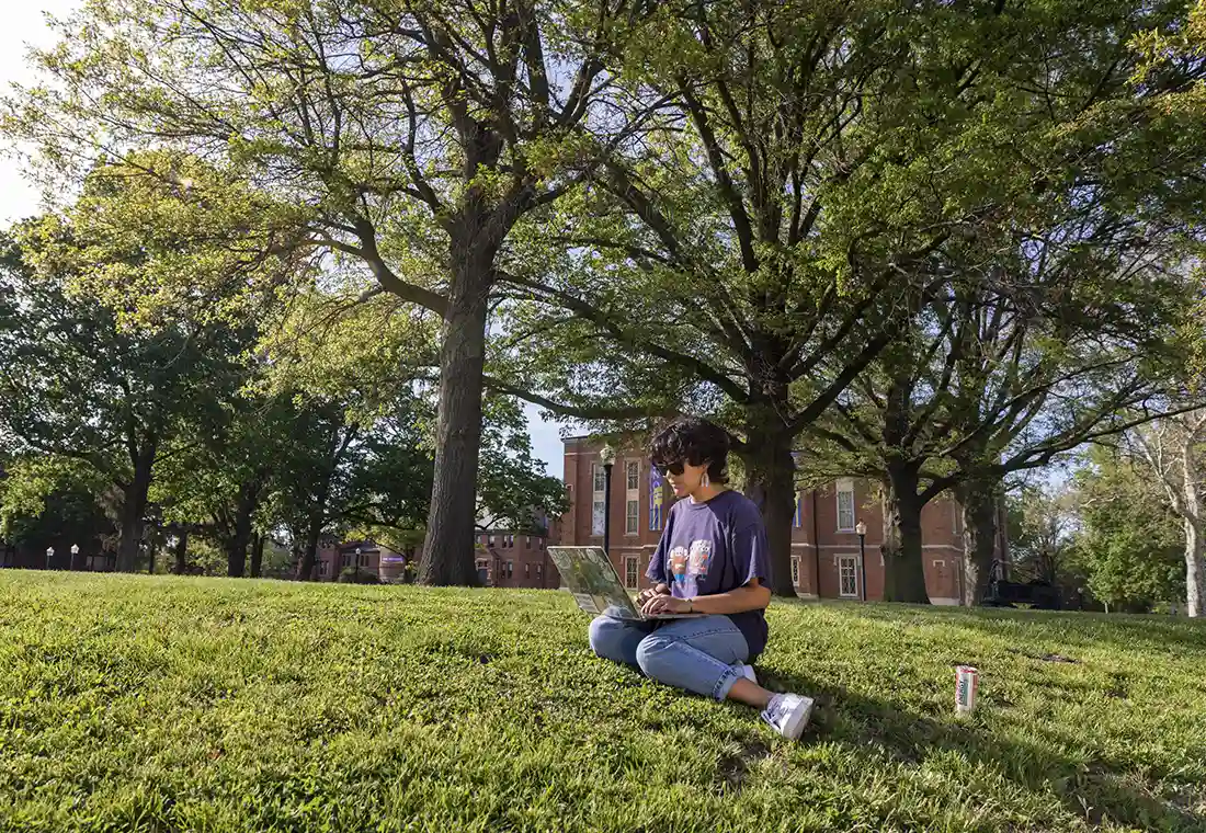A student with a laptop sits on the campus lawn on a sunny day at Knox College.
