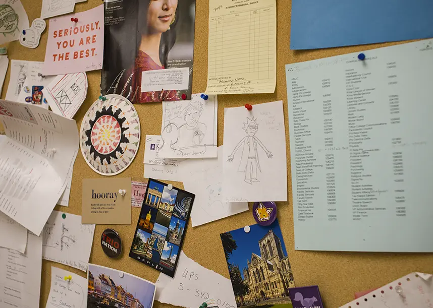 A bulletin board outside of the mailroom that has different flyers and drawings posted to it.