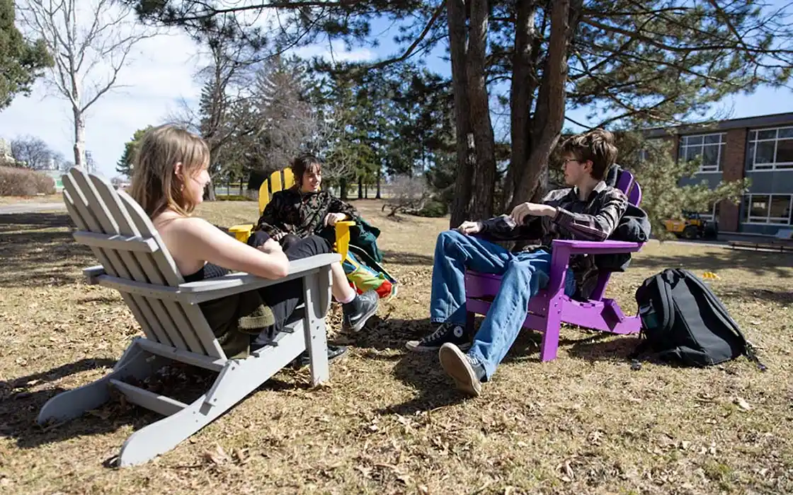 Three students sit in Adirondack chairs on campus, smiling and talking.