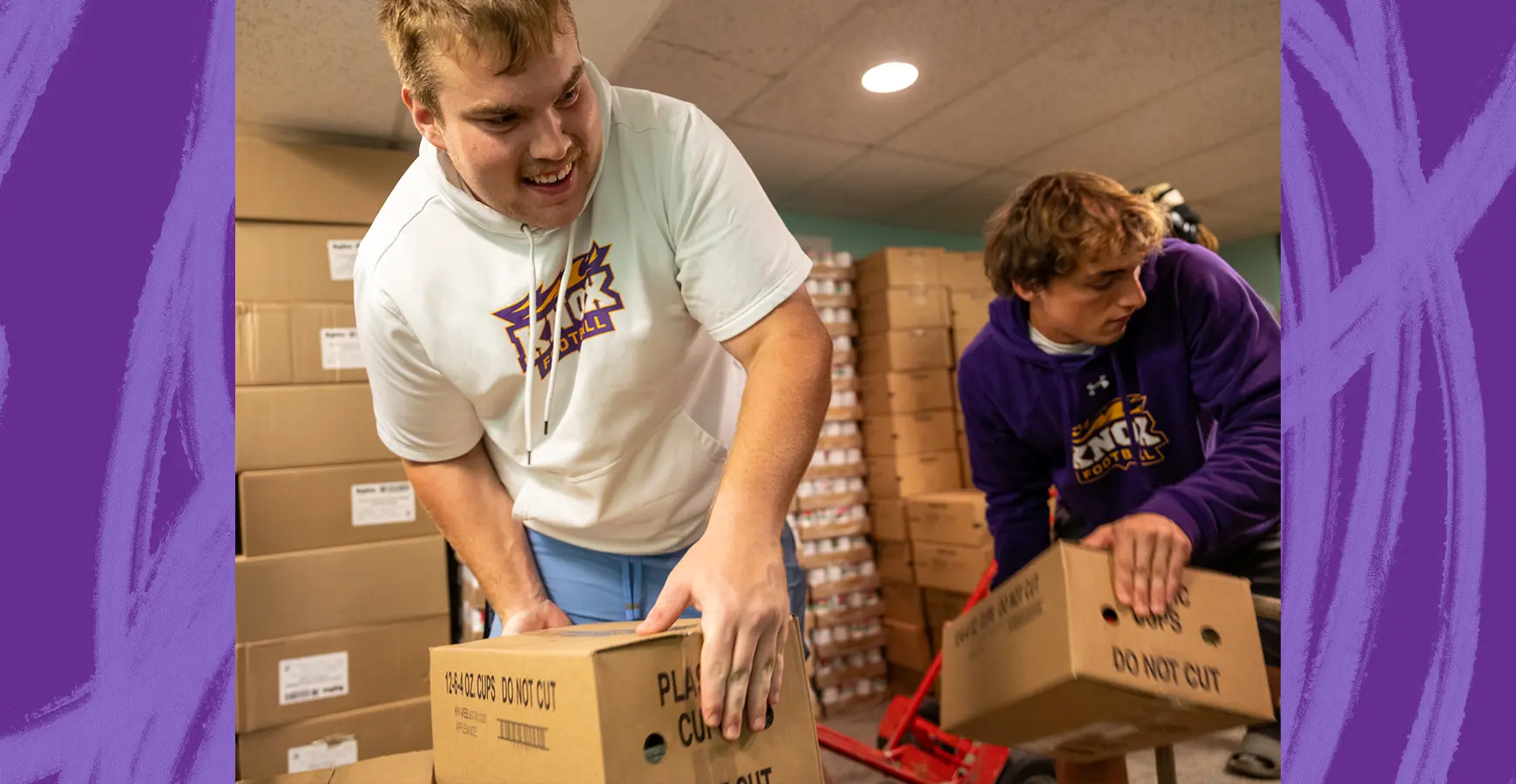 Two students lift cardboard boxes in a storage area with many other boxes stacked around them.