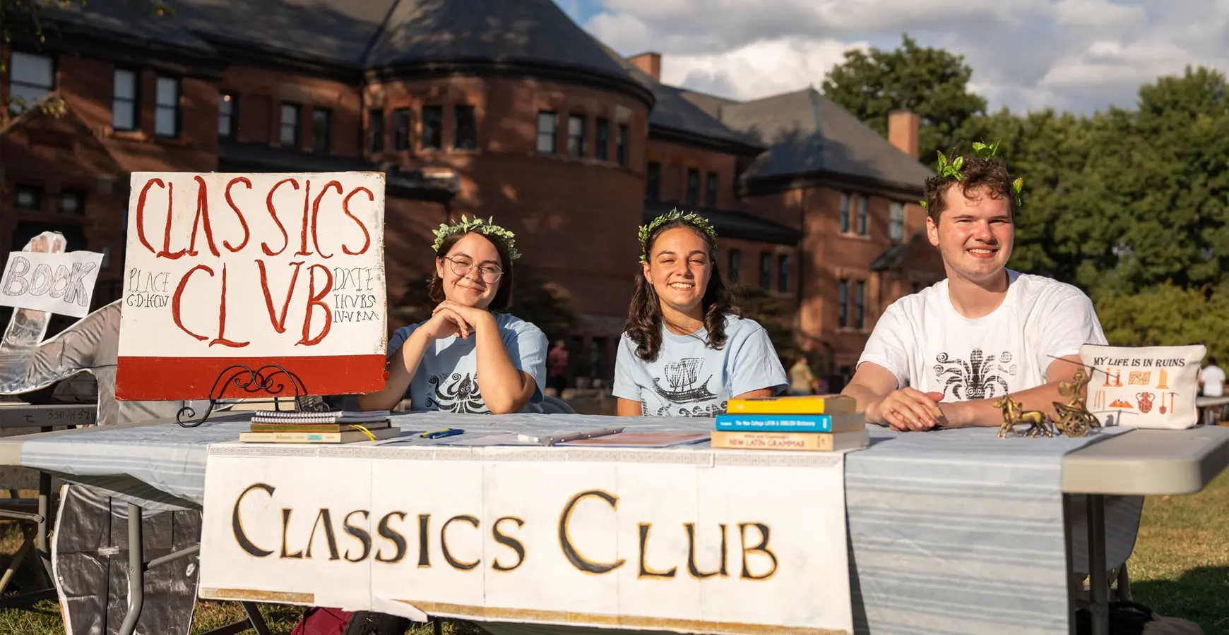 Three students sit at a table outside with a sign that says Classics Club