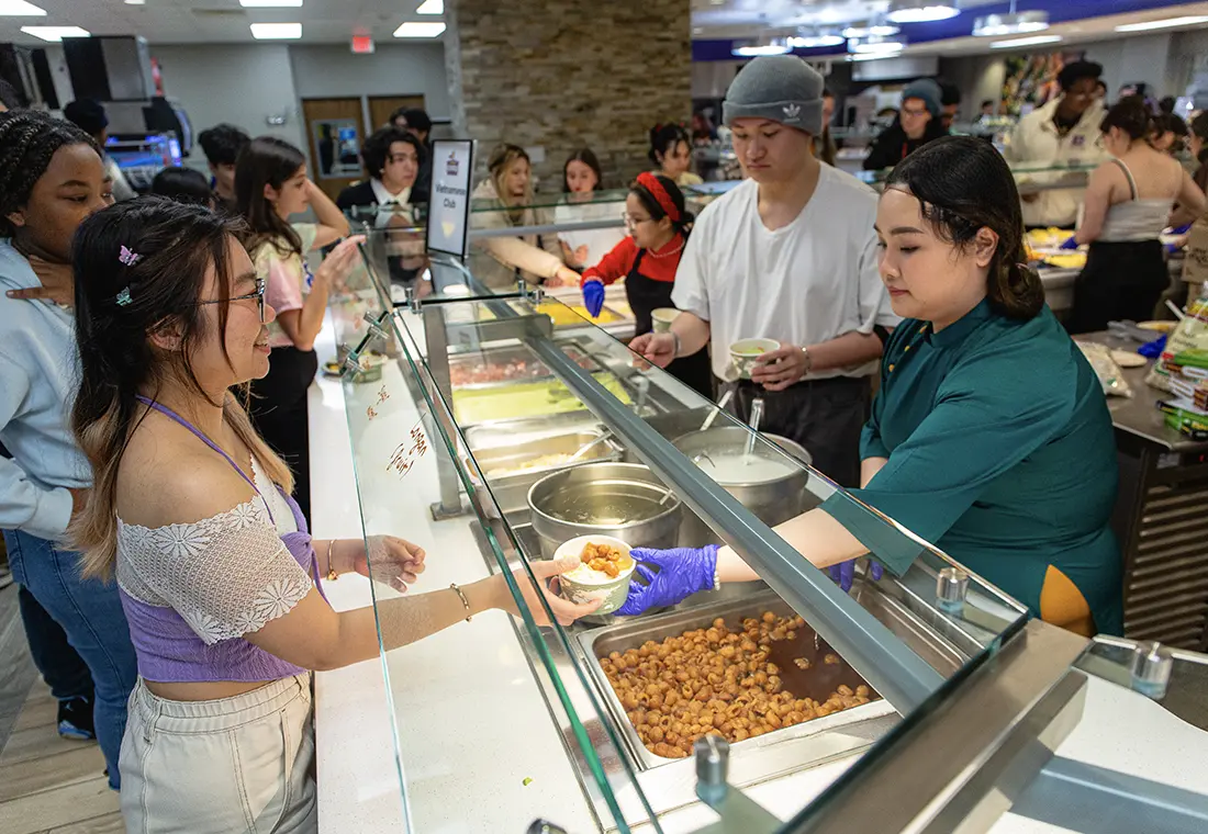 Students stand in a line, having food served to them in the cafeteria.