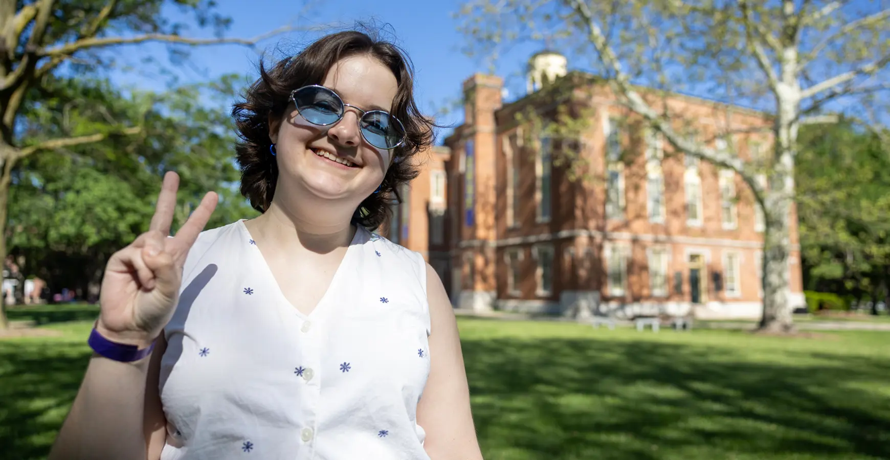 A student wearing blue tinted glasses smiles at the camera and holds up two fingers as a peace sign. Old Main is in the background. 