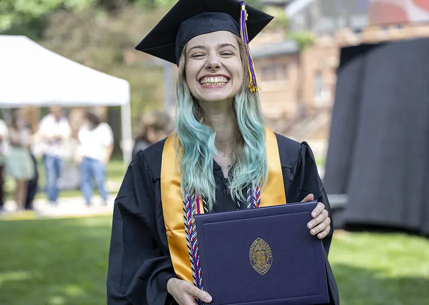 A Knox graduate with long blue hari in cap and gown smiles at the camera and holds a Knox College diploma.