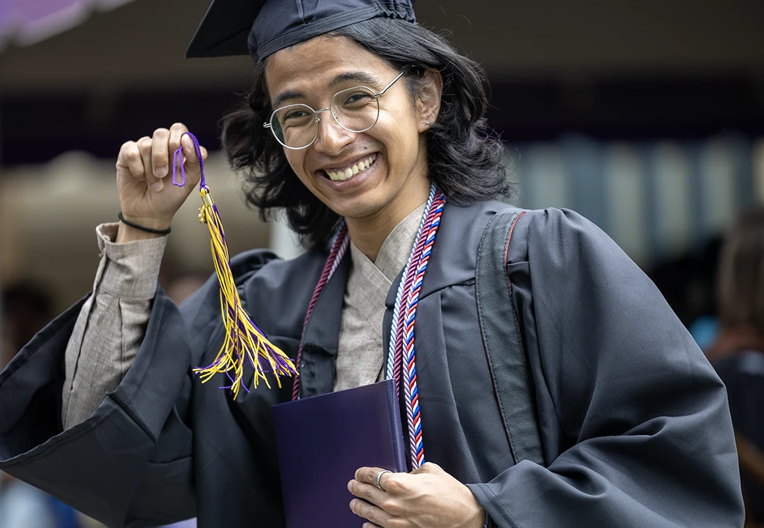 A graduating student in cap and gown smiles at the camera holding a tassel in one hand and a diploma in the other.