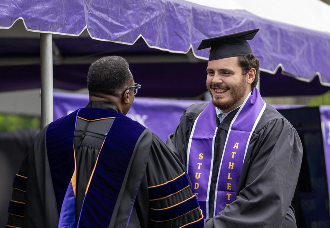A graduating student in cap and gown shakes President C. Andrew McGadney's hand. The student wears a student athlete stole.