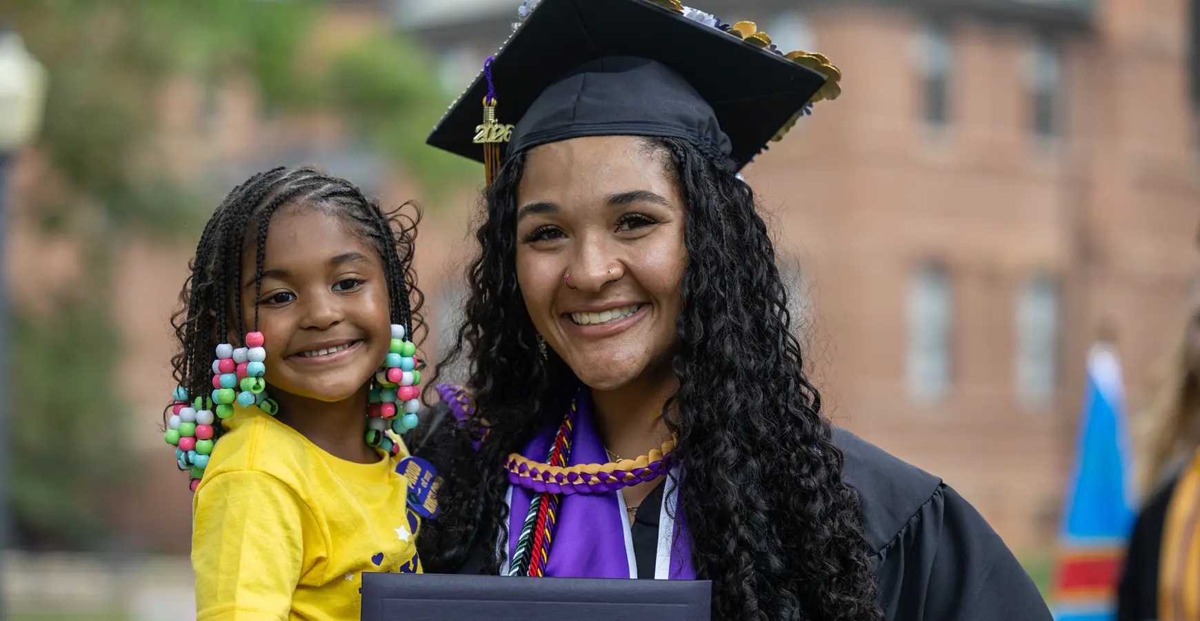 A graduate smiles at the camera, wearing academic regalia and holding a child, who also smiles.