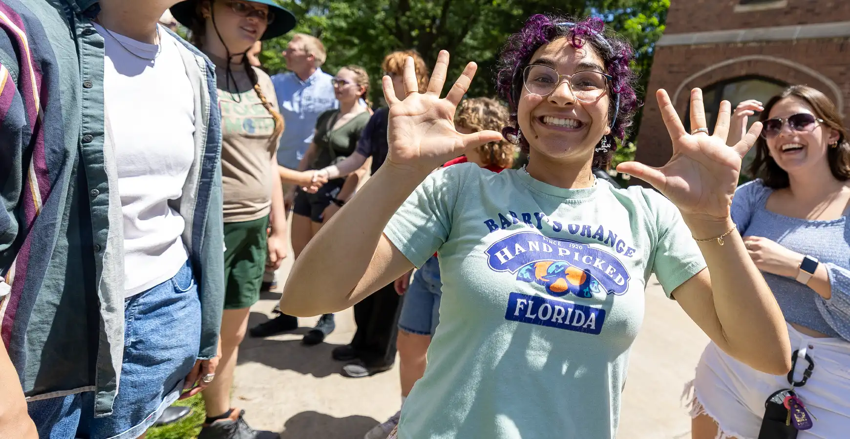 A student smiles at the camera while holding up jazz hands. Several students are visible around her, also smiling.