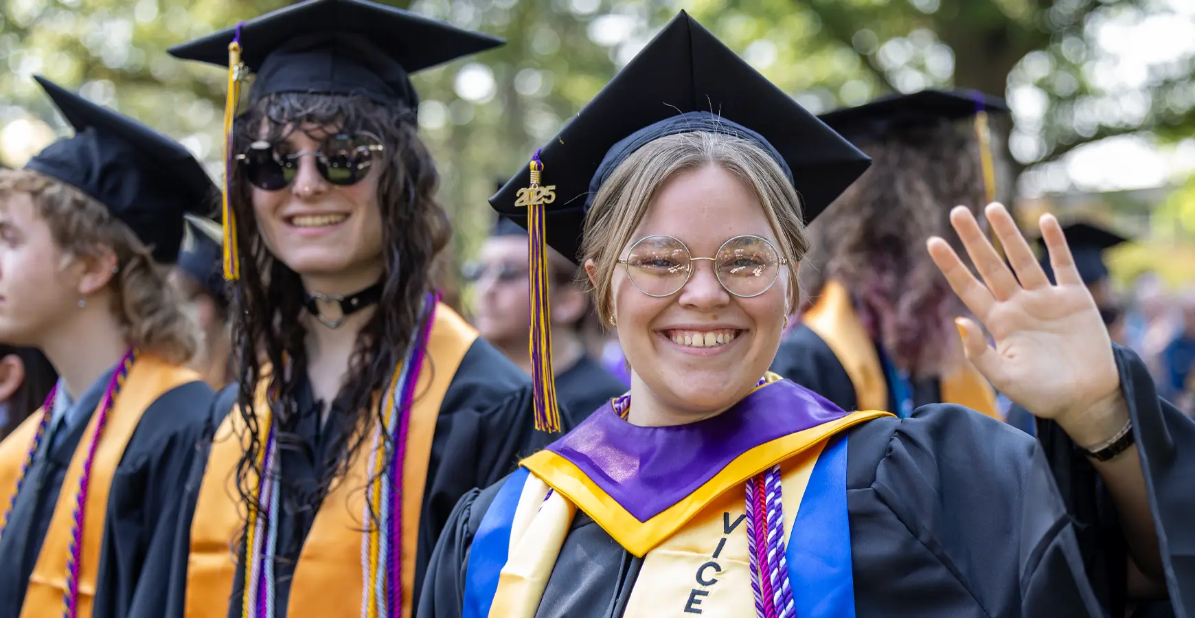 Two Knox graduates in caps and gowns with purple and gold stohls smile and wave at the camera. 