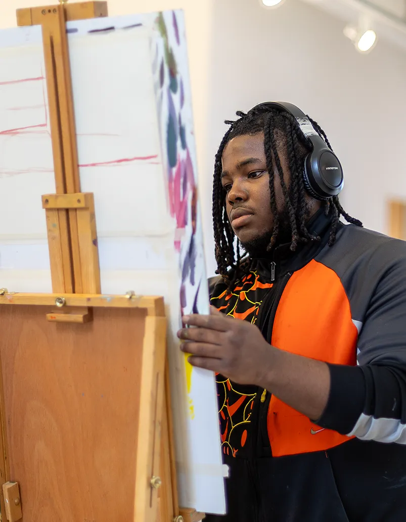 Wearing headphones, a student works intently on a canvas.