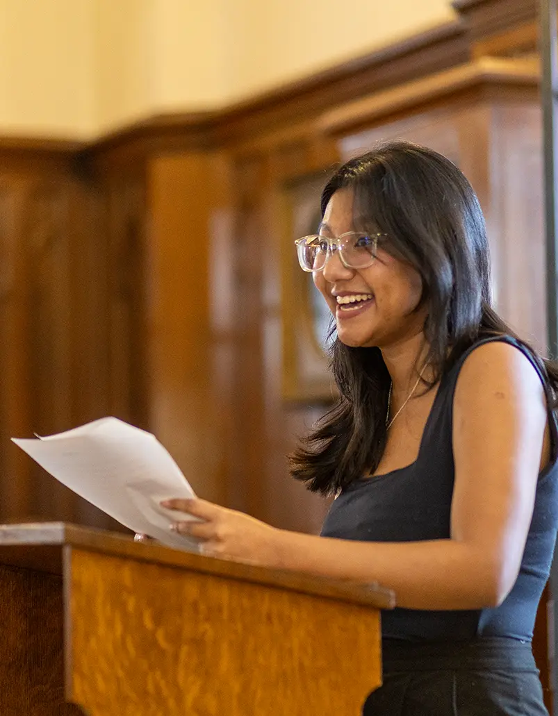 A smiling student standing a podium holding a manuscript.