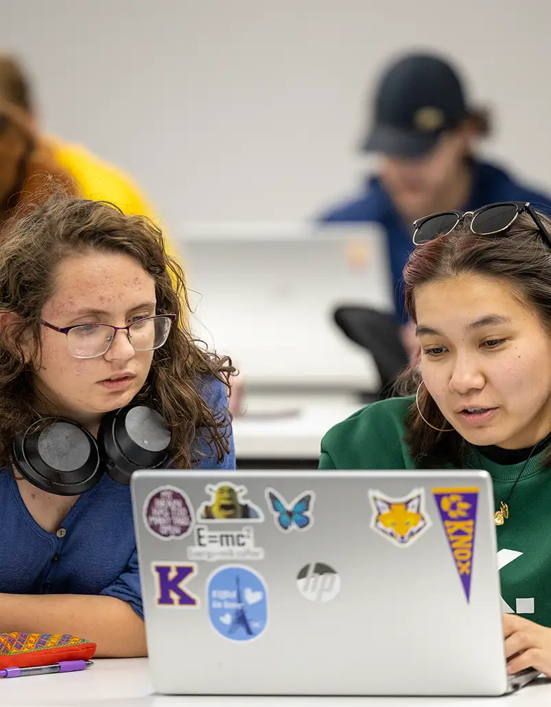 Two students looking at a laptop, stickers are on the front of the computer. 