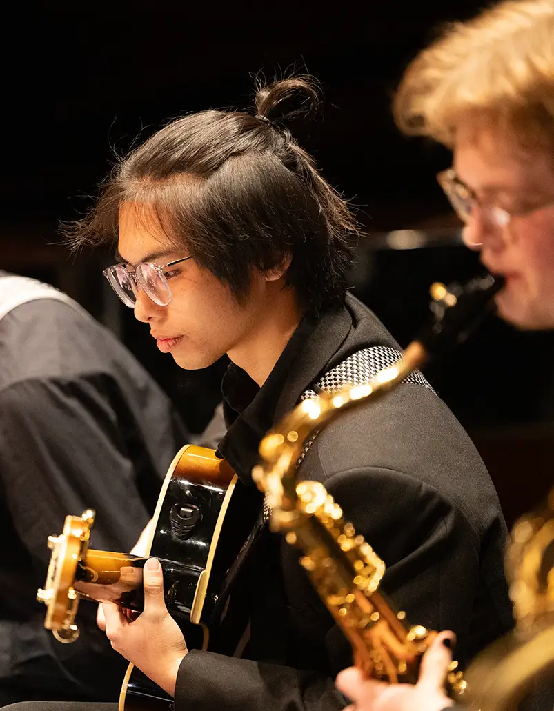 Two Knox students dressed in performance black play their instruments during a performance. One plays guitar, one plays saxophone.