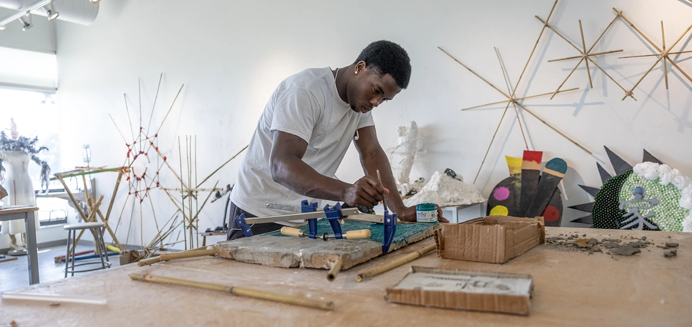 In an art studio, a student leans over an art project as he works on it. He's surrounded by other art projects.