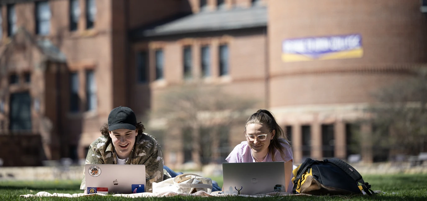 Two students lie on the ground in front of Alumni Hall, studying with their laptops open.