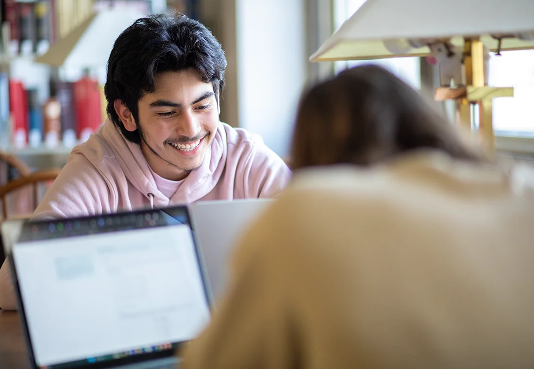 Two students sit in front of laptops at a study table in Seymour Library; one student faces the camera smiling, another student's back is to the camera.