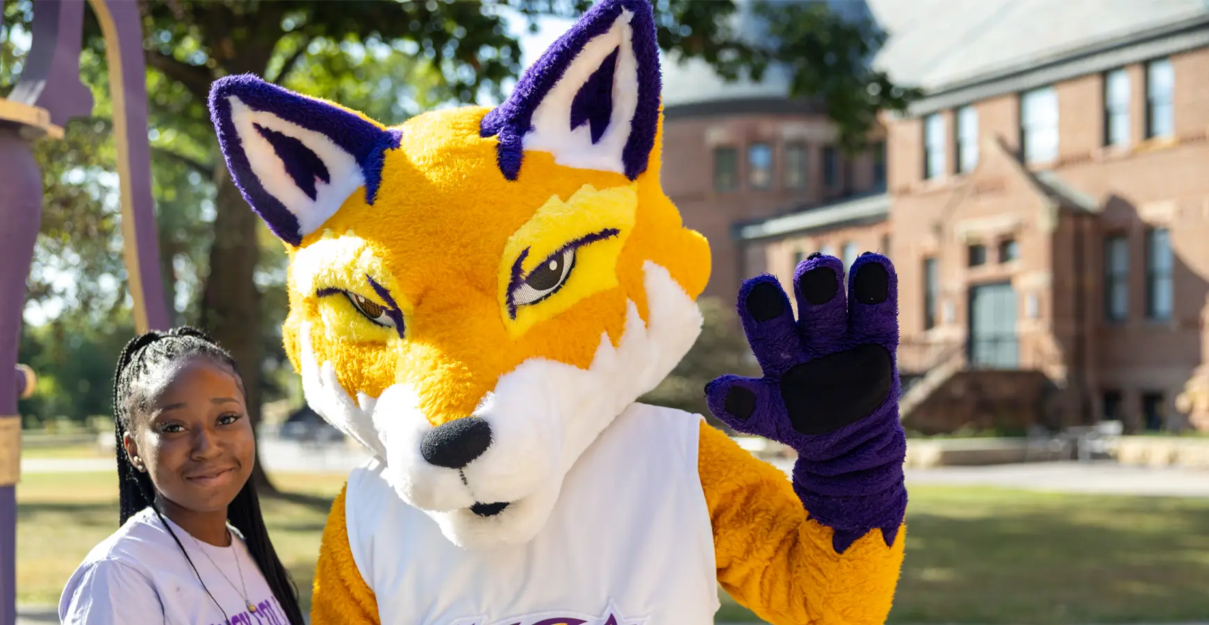 A student stands next to Blaze and smiles. Blaze waves at the camera. 