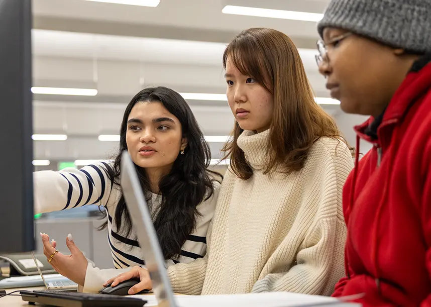 Three students gathered around a computer screen. One is pointing pointing at the screen.