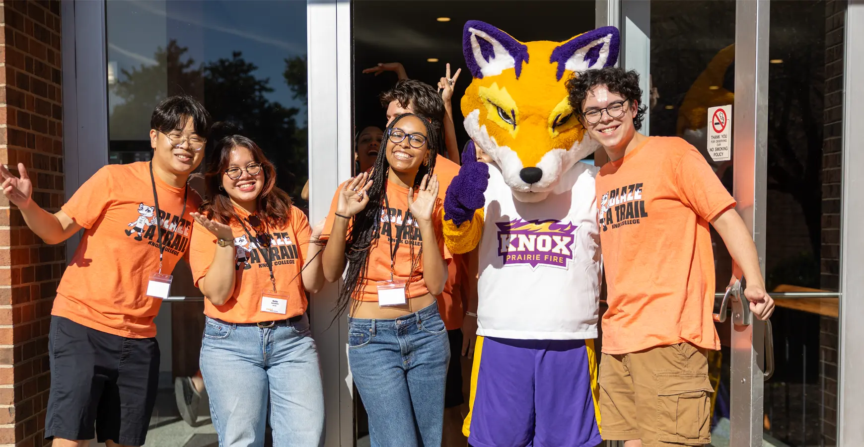 A group of students standing with Blaze the Fox outside waving and smiling at the camera.