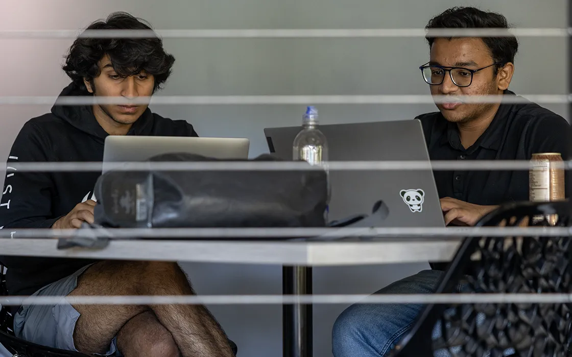 Two students with laptops studying at a round table are viewed through blinds on a windwo.