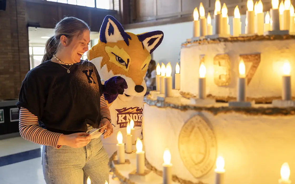 A Knox student and Blaze, the Knox Prairie Fire fox mascot, stand beside a large replica of a cake that's light by candle lights. 