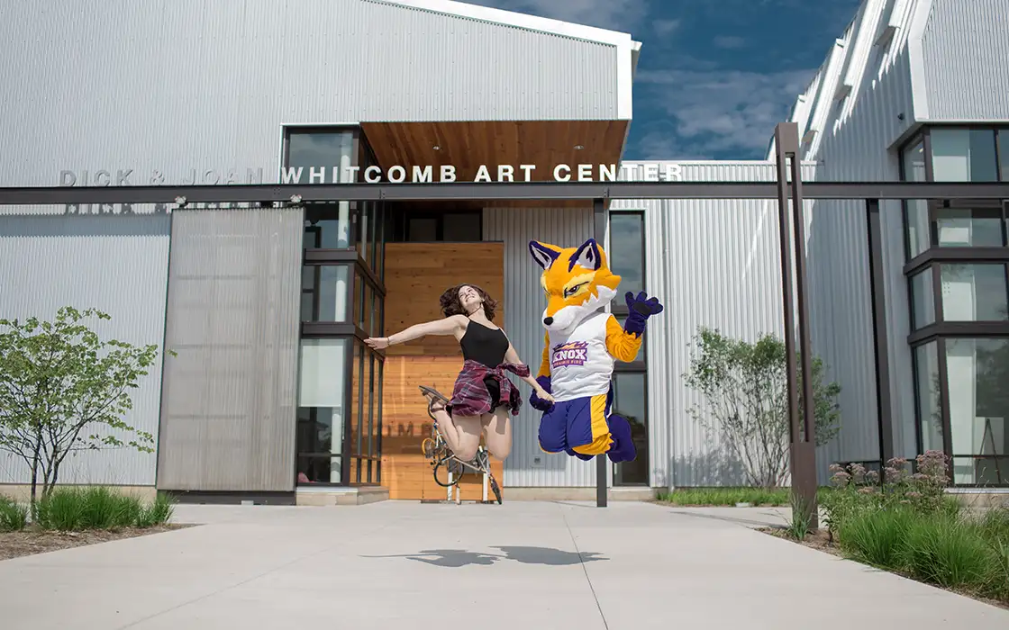 A student and Blaze jump in the air in front of the Whitcomb Art Center.