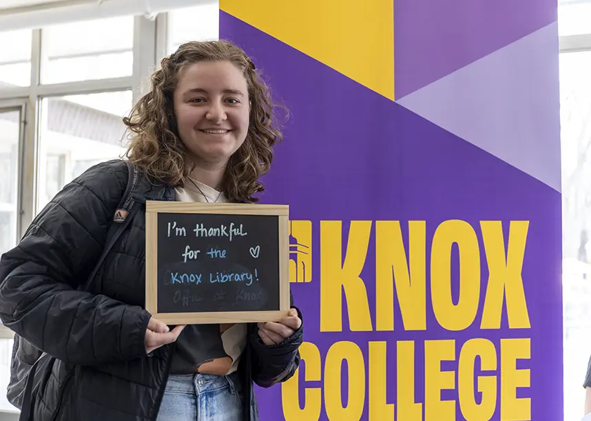 A student stands next to a Knox College banner holding a chalkboard that says I'm thankful for the Knox library!