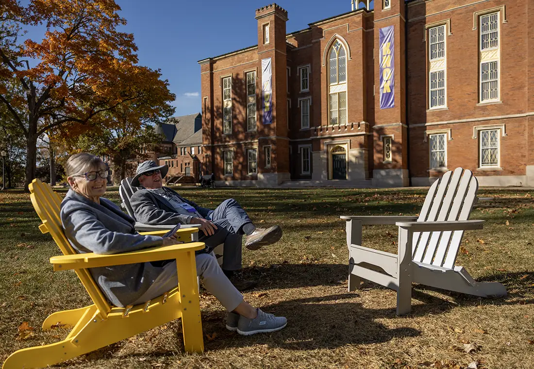 Two Knox alumni sit in yellow and purple adorondak chairs on the lawn in behind Old Main.