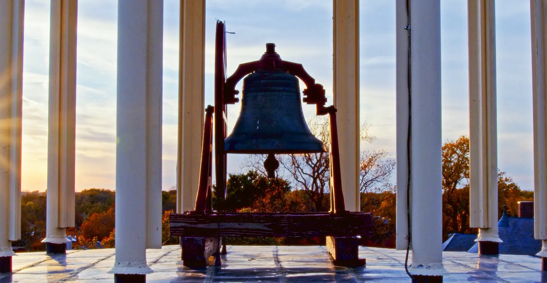 A close-up of the Old Main bell with the tops of campus trees at sunset in the background.