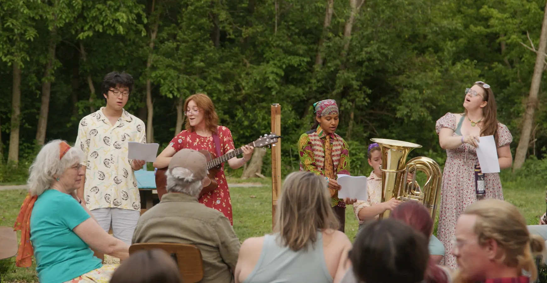 A group of students performs music outdoors in a wooded park, singing and playing instruments including a guitar and tuba for a seated audience.
