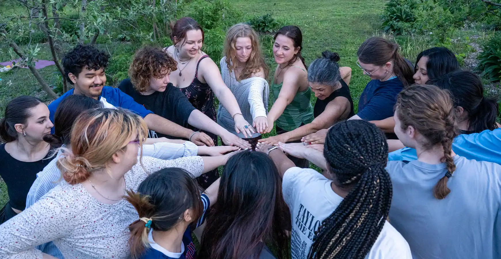A large group of individuals stand in a circle; all of them extend their hands to the middle of the circle.