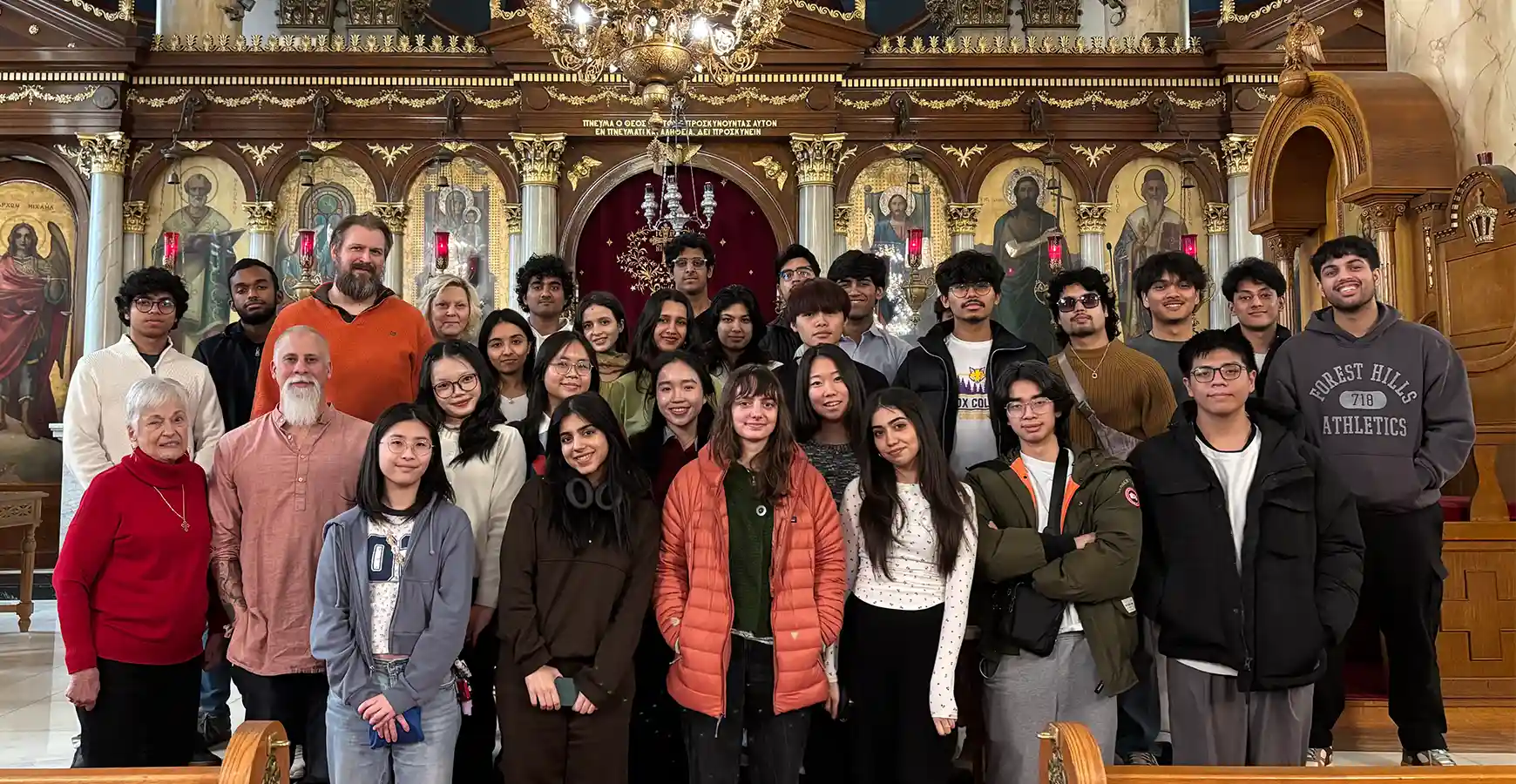 A large group of 25 plus individuals stands together, smiling at the camera. The backdrop shows historic frescos within arches, depicting religious figures.  