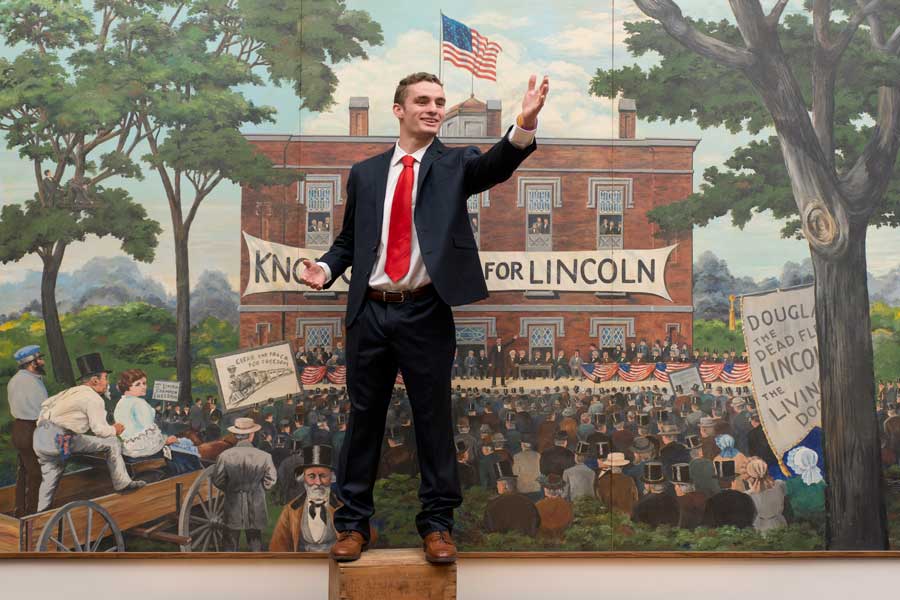Charles Harned '16 poses in front of a Lincoln mural in Knox College's Alumni Hall. 