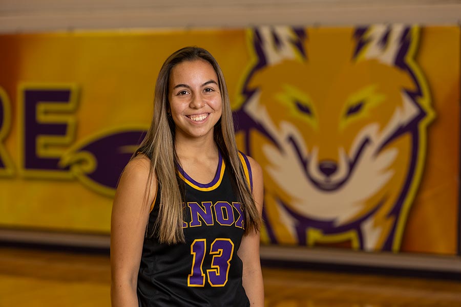 Saxon stands on the basketball court with a black basketball uniform with 13 in purple letters and a yellow border. The Knox mascot, Blaze, an orange-yellow fox is presented on a large wall poster behind her. 