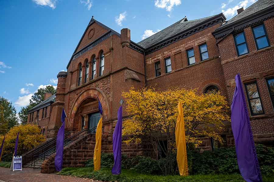 Exterior view of Alumni Hall, a red brick building, with purple and gold flags in front