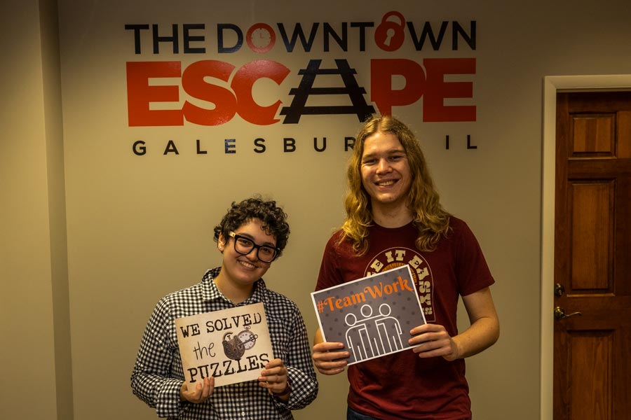 In front of beige wall that has The Downtown Escape signage printed above two people, Maria and Oscar, holding signs that say "We Solved the Puzzles" and "Team Work." 