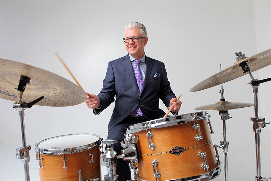 Drummer, Matt Wilson, sits in front of a modern, fire-orange drum set in a dark blue suit and purple tie holding a drum stick in right hand above drum cymbal.