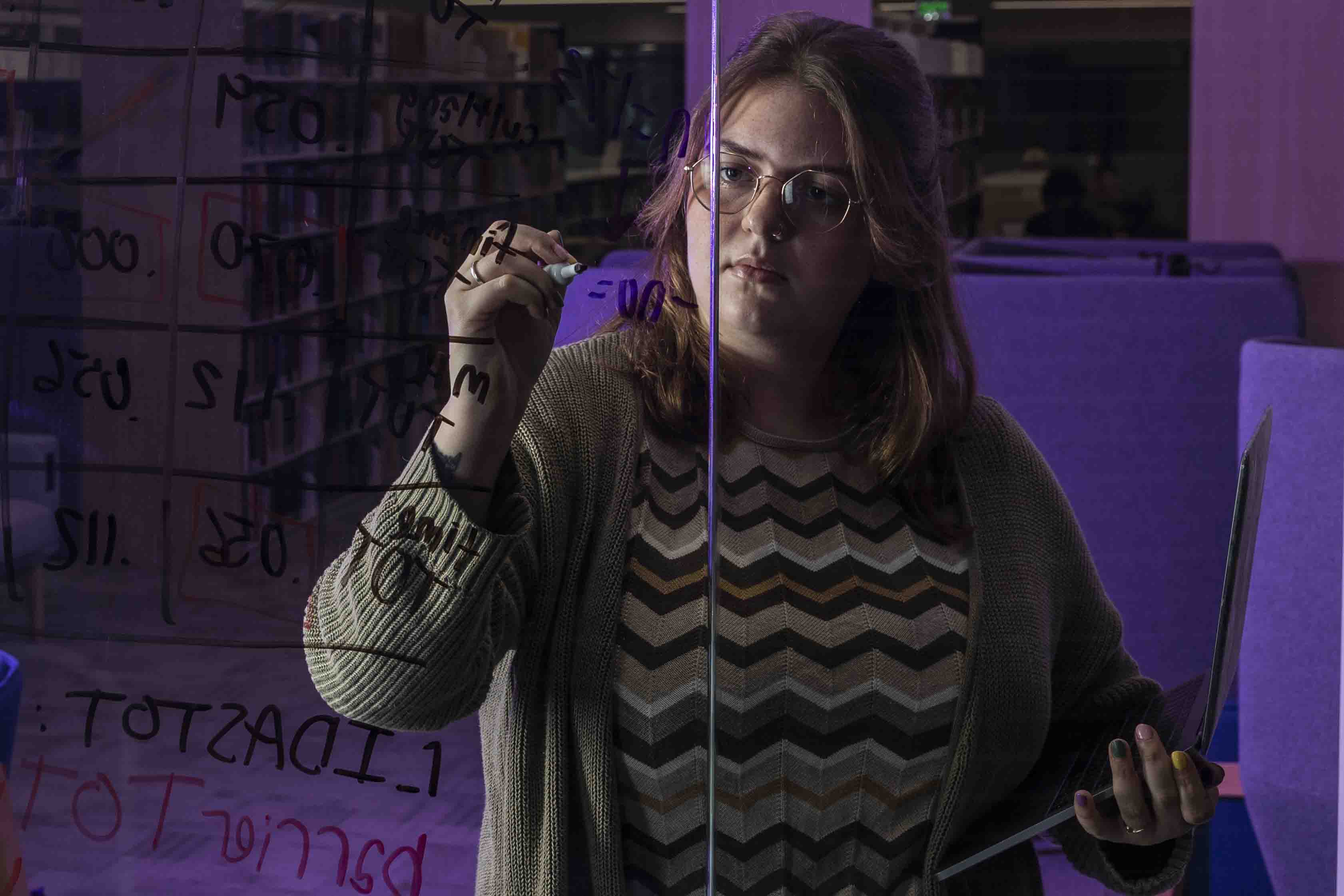 Maddie stands behind a clear class board with black marker in her hand writing down formulas in a darkened, purple lit background.