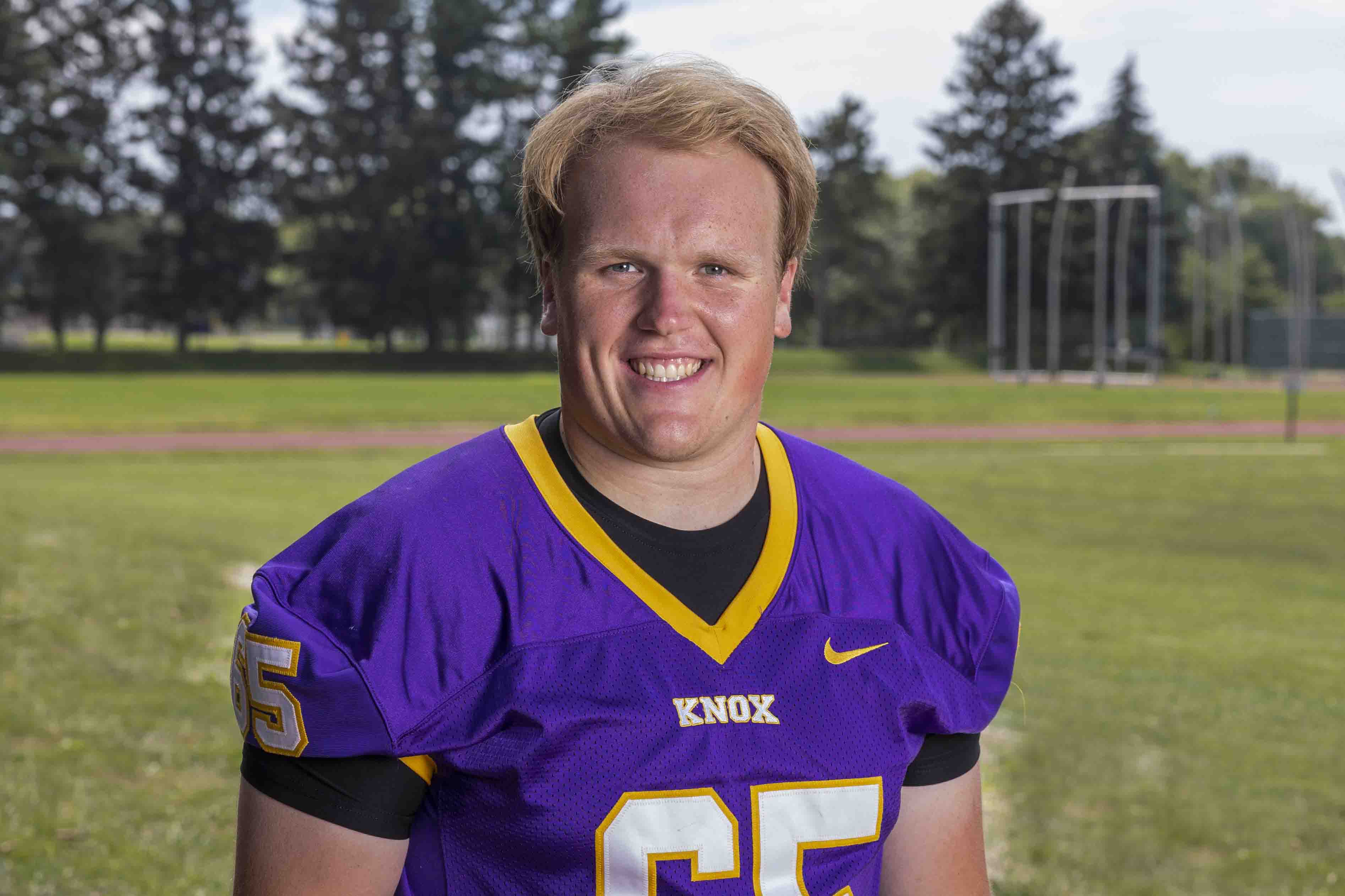 Charlie stands with a purple football jersey in front of green, grassy field. 