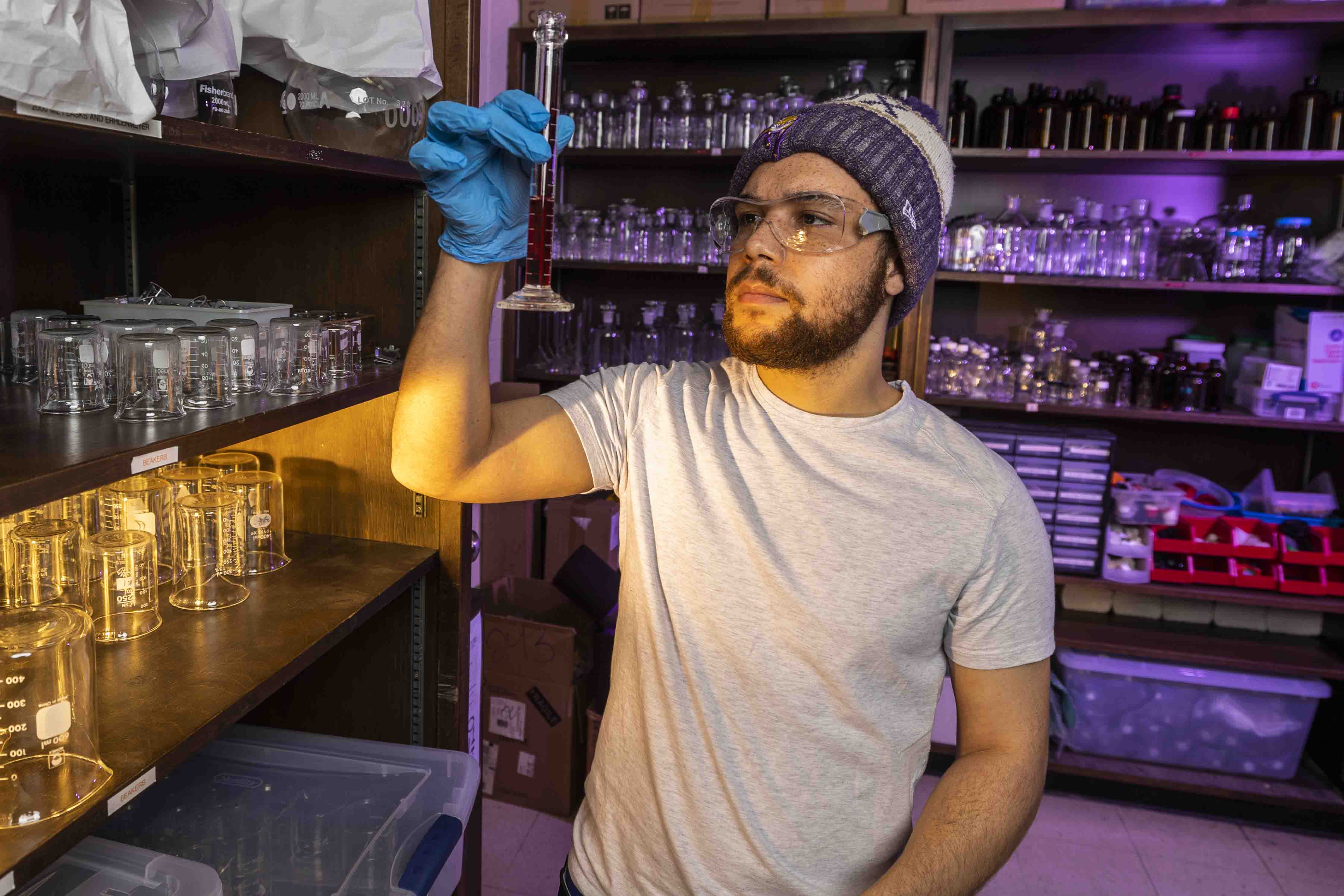 John, in a white t-shirt and purple Knox beanie and wearing blue surgical gloves, holds up and observes a measuring cylinder. 