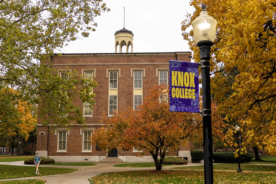 Old Main is pictured in early fall. A lightpost is in the foreground with a banner that says Knox College.