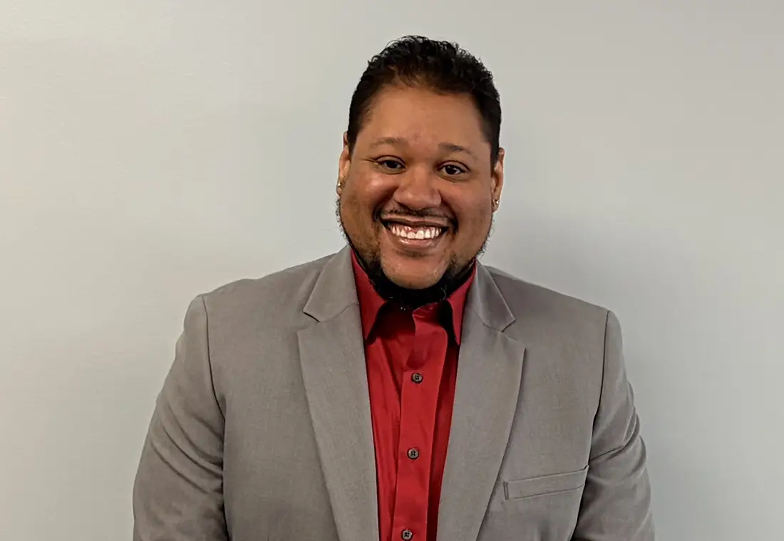 DeAndre Henderson ’10 in a grey suit with a red shirt standing in front of a blank wall.