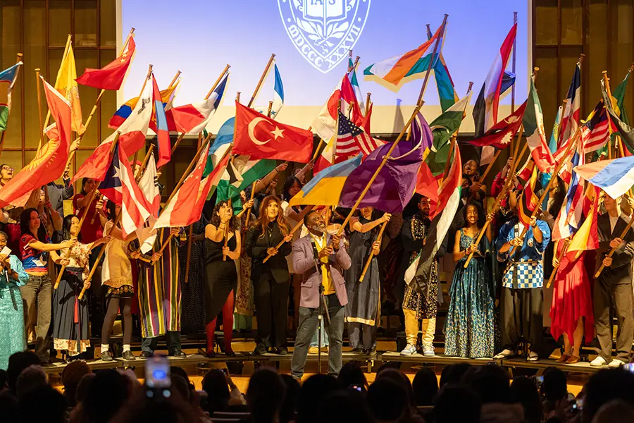 A large group of Knox students stands on a stage with President C. Andrew McGadney waving flags from several different countries around the world.